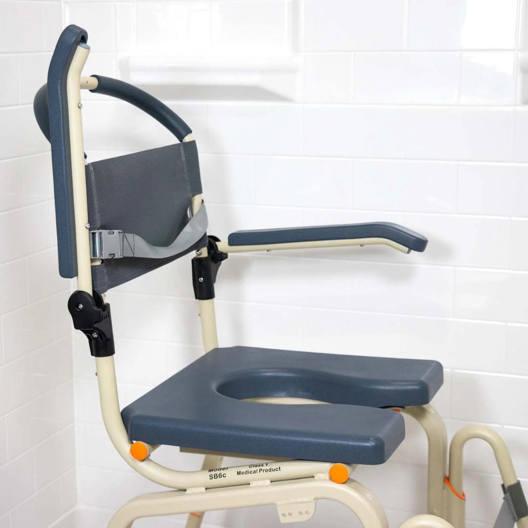 The ShowerBuddy SB6C Roll-in shower chair commode, featuring armrests, an open seat, sturdy metal frame, and dark gray padded seat, backrest, and armrests for comfort, is positioned against a white tiled wall.