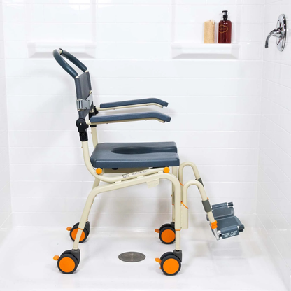 A shower with white tiled walls features the ShowerBuddy SB6C Roll-in Shower Chair Commode, equipped with wheels, armrests, and footrests for mobility assistance. Shampoo and soap bottles rest on a built-in shelf.
