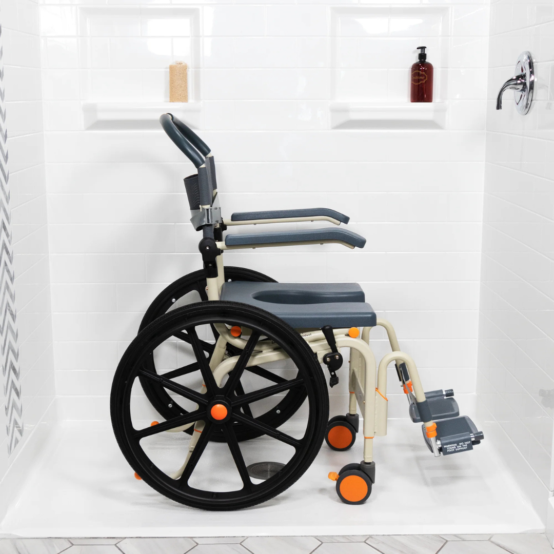 The ShowerBuddy SB6W Roll-In Shower Wheelchair with black wheels and orange accents is placed in a white tiled roll-in shower with a handheld showerhead and shelves holding bottles.