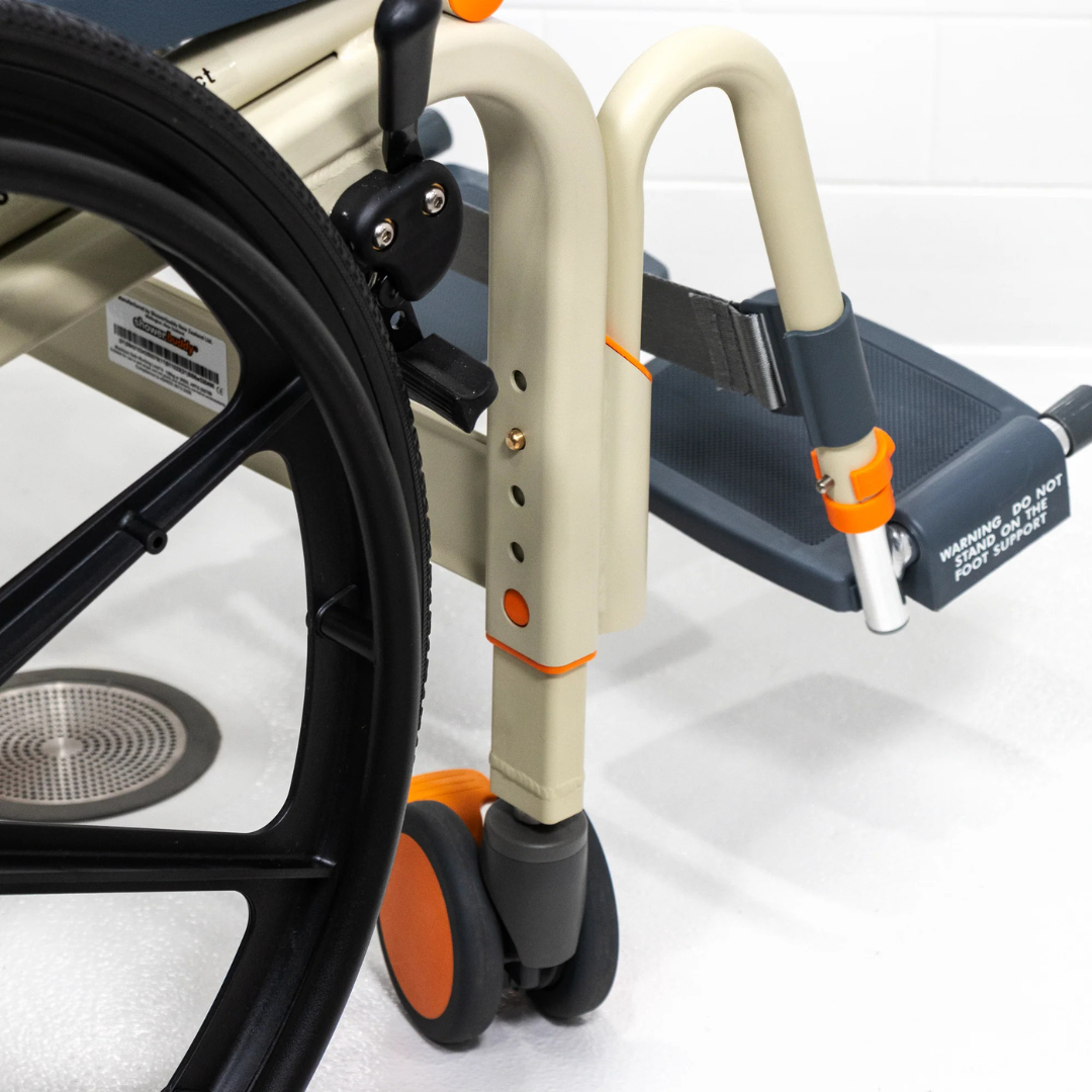 Close-up of the ShowerBuddy SB6W Roll-In Shower Wheelchair’s front section, showing the front wheel, footrest, orange accents, and seat against a white floor and tiled wall—ideal for accessible roll-in showers.