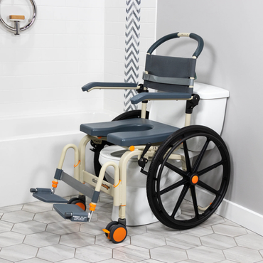 The ShowerBuddy SB6W Roll-In Shower Wheelchair, in gray and cream, features a commode seat, large rear wheels, foldable armrests, and footrests. It is positioned over a toilet in a tiled bathroom for easy transfers to a roll-in shower.