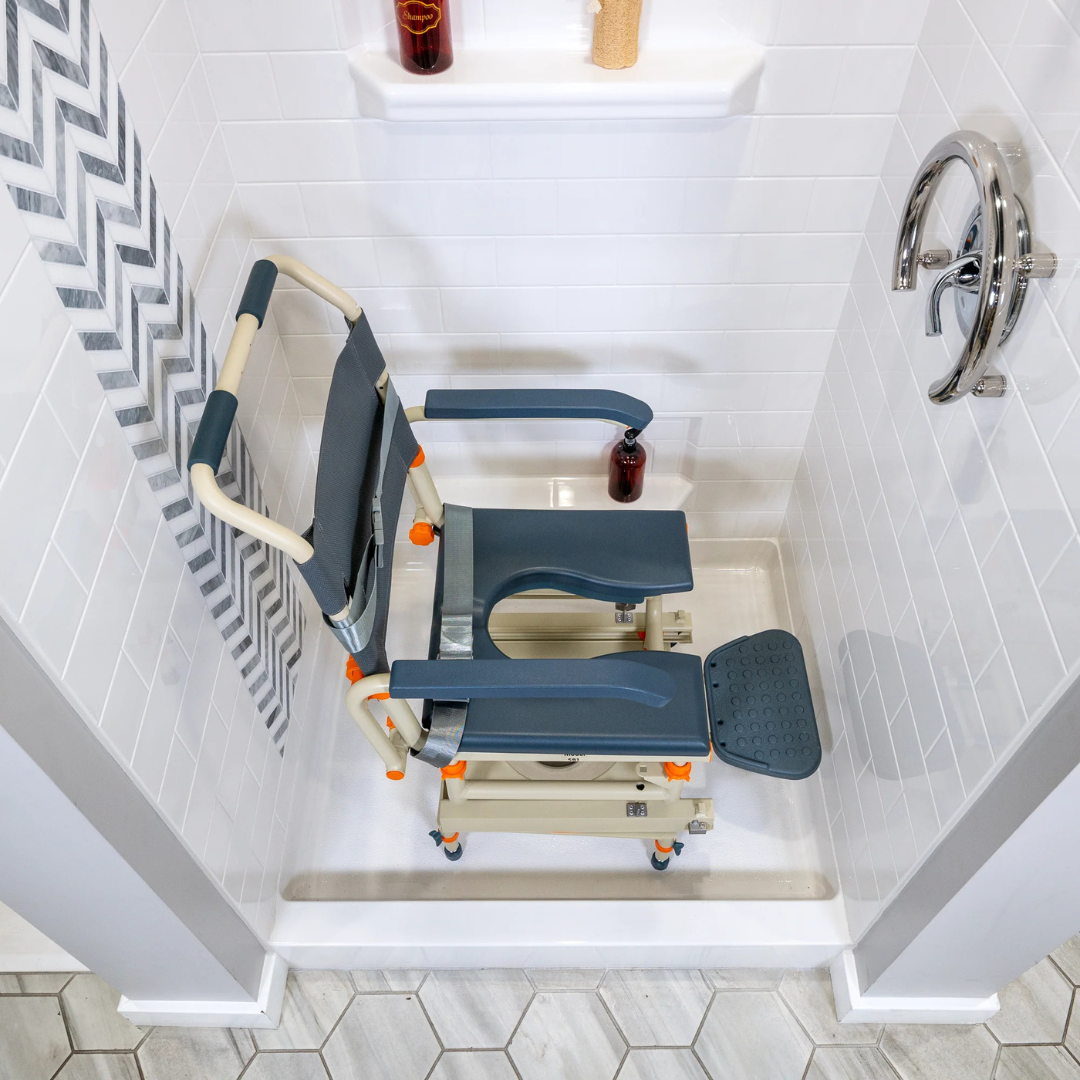 A ShowerBuddy® SB1 Transfer System with Rolling Shower Chair, featuring armrests, a padded seat with opening, and footrests, is placed inside a white-tiled shower with a striped curtain and chrome handle.