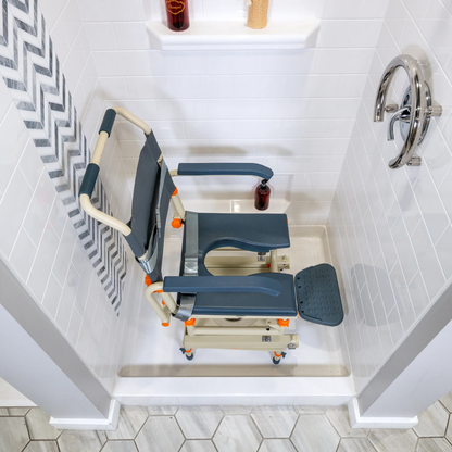 A ShowerBuddy® SB1 Transfer System with Rolling Shower Chair, featuring armrests, a padded seat with opening, and footrests, is placed inside a white-tiled shower with a striped curtain and chrome handle.