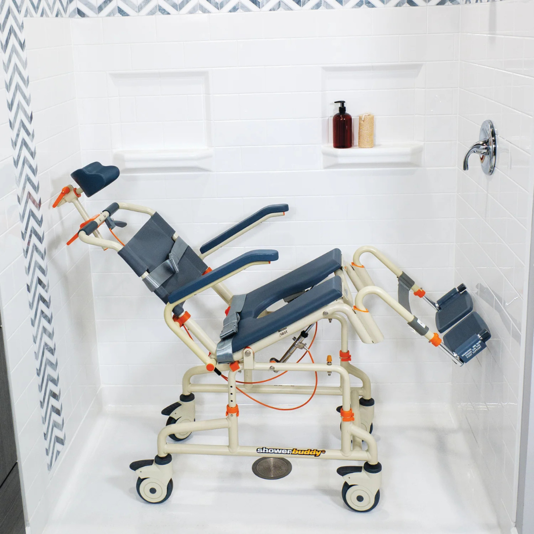 The ShowerBuddy SB3T Roll-In Shower Chair with Tilt Recline, featuring wheels, adjustable backrest, armrests, and footrests, is positioned inside a tiled shower stall. Soap and shampoo bottles sit on a built-in shelf.