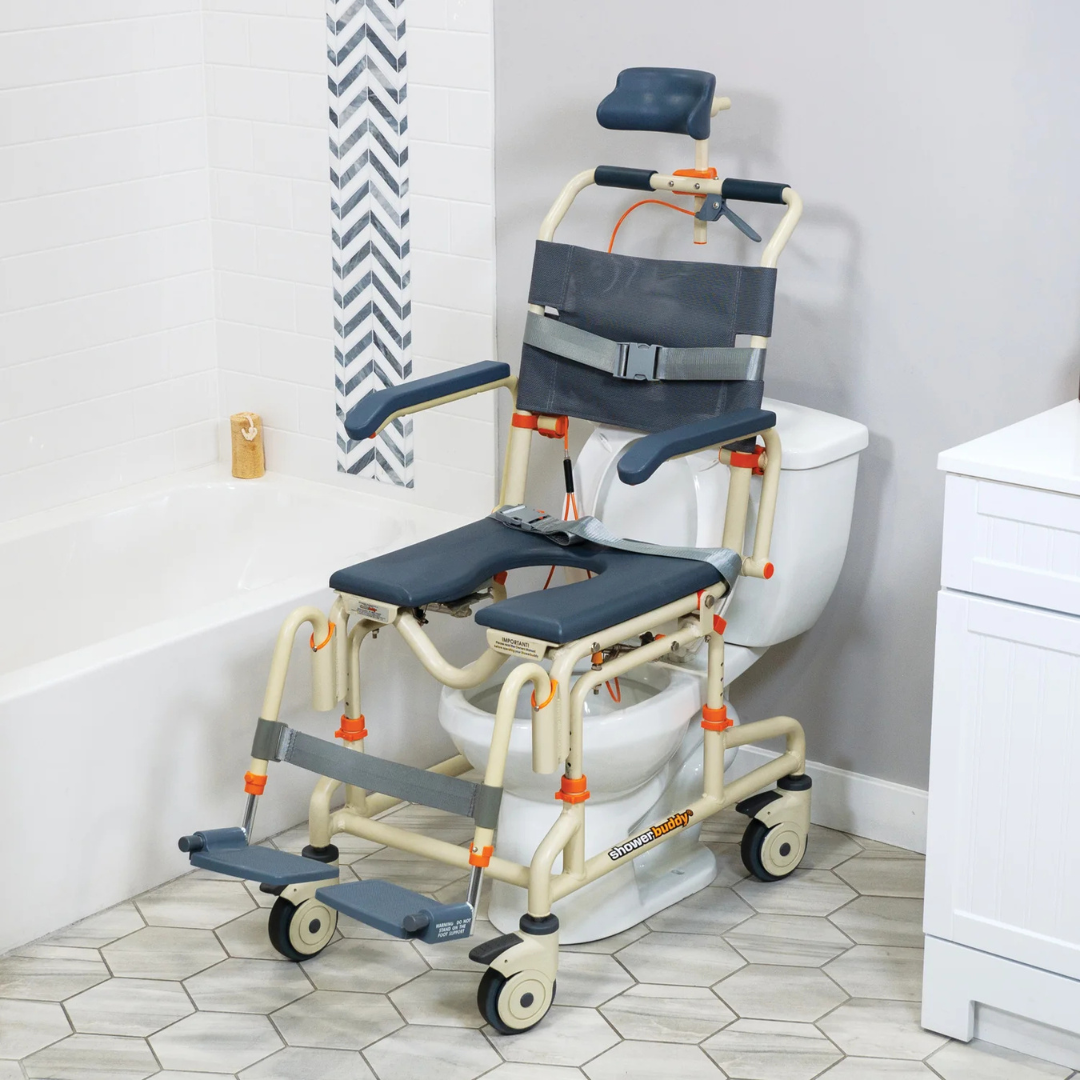 The ShowerBuddy SB3T Roll-In Shower Chair with Tilt Recline, featuring armrests, headrest, seatbelt, and footrests in beige and gray, is positioned over a white toilet in a bathroom with hexagonal floor tiles and white tiled walls.