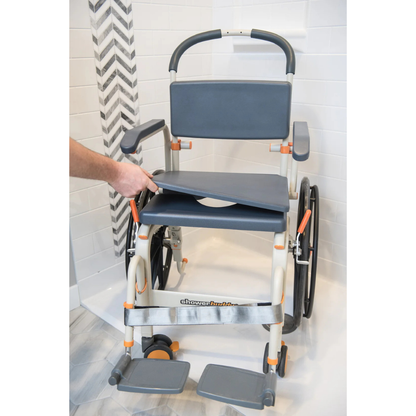 A person lifts the padded seat of a ShowerBuddy SB6W Roll-In Shower Wheelchair, featuring footrests, armrests, and large self-propelled rear wheels, in a tiled bathroom next to a roll-in shower.