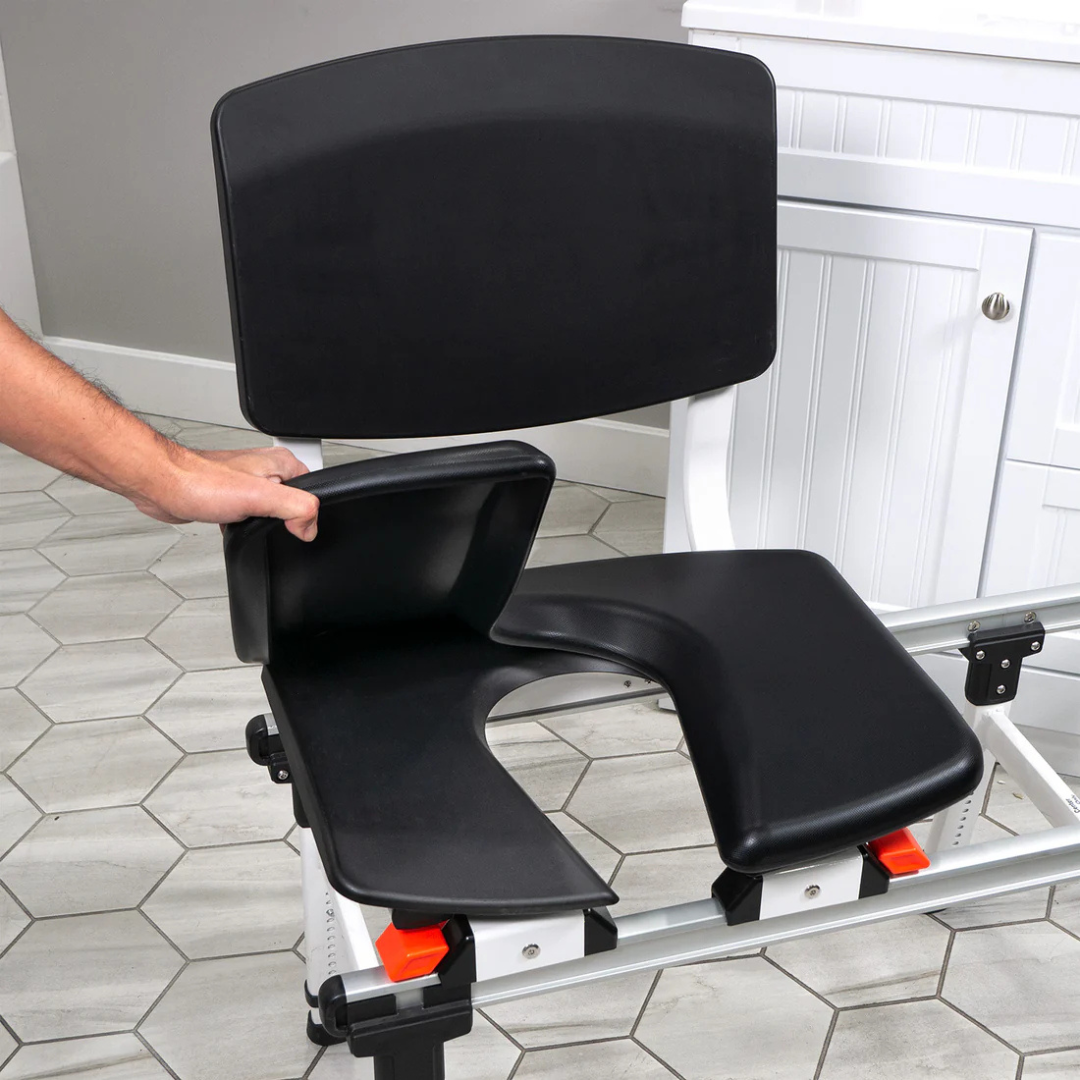 A person lifts a cushioned seat cover, an accessory for ShowerGlyde® Transfer Benches, on a black commode chair with a U-shaped hard-plastic seat, enhancing bathing comfort in the bathroom.