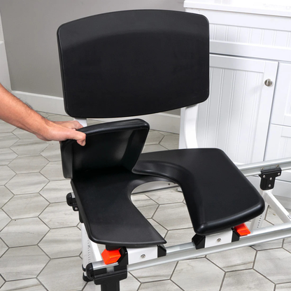 A person lifts a cushioned seat cover, an accessory for ShowerGlyde® Transfer Benches, on a black commode chair with a U-shaped hard-plastic seat, enhancing bathing comfort in the bathroom.