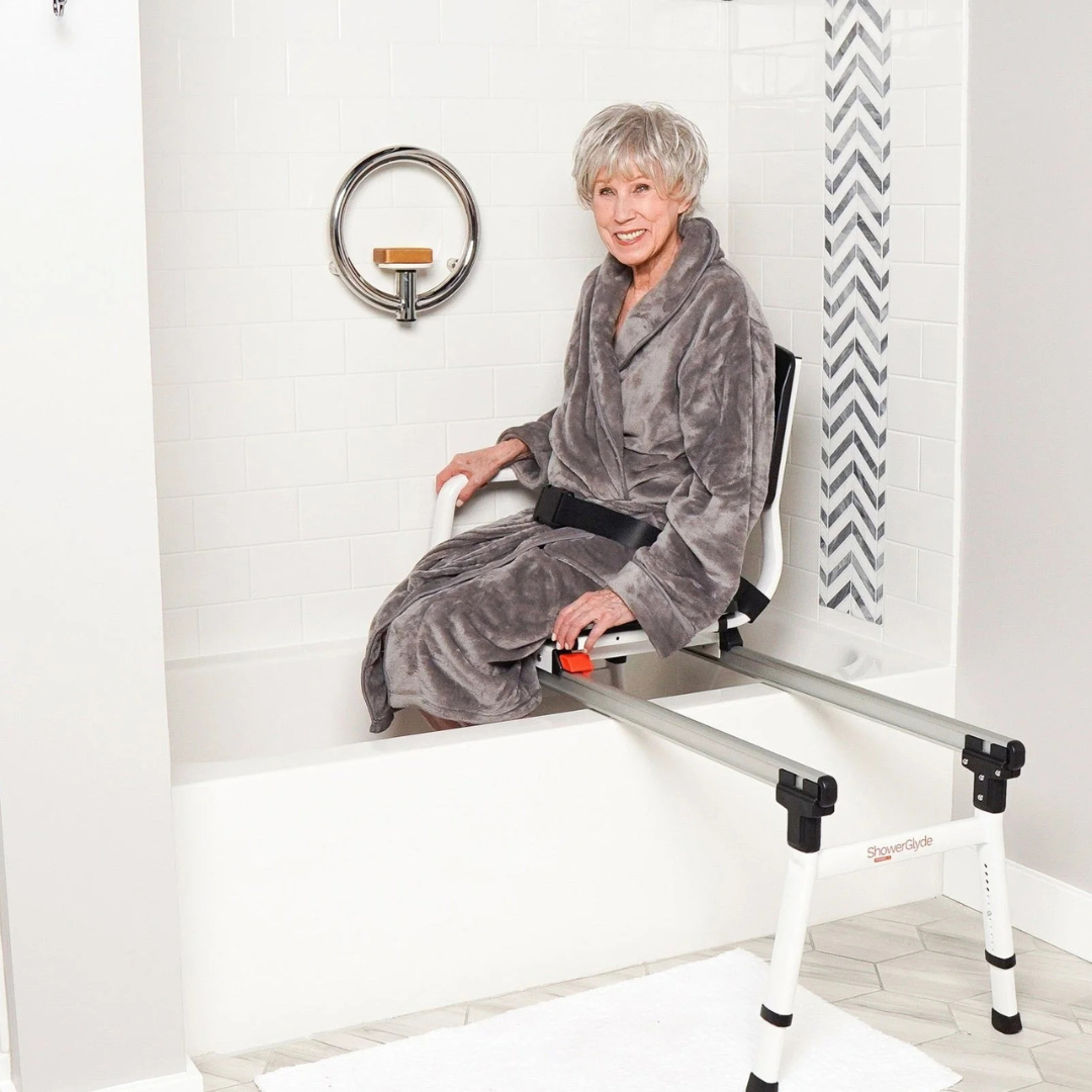 An older woman smiles as she sits safely on the ShowerGlyde SG1 Bathtub Sliding Transfer Bench, which bridges the tub edge in a white-tiled bathroom with a towel ring and chevron accent tiles.