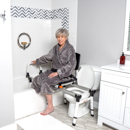 An older woman in a gray robe uses the ShowerGlyde SG2 Sliding Transfer Bench to safely move from the toilet to the adjacent bathtub in a modern, accessible bathroom, smiling and gripping the safety handles for support.