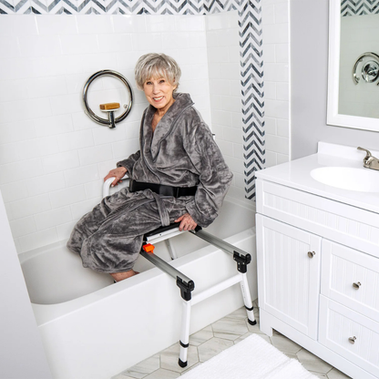 An older woman in a gray bathrobe uses the ShowerGlyde SG3 Sliding Transfer Bench with Swivel Seat to safely enter a white bathtub in a modern bathroom with white cabinets and tiled walls.