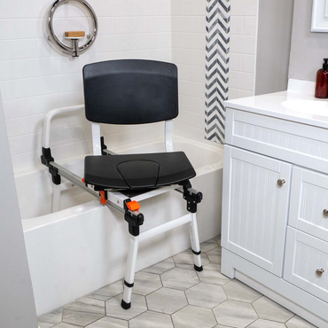 The ShowerGlyde SG3 Sliding Transfer Bench with Swivel Seat in black and white is placed over a bathtub edge near a white vanity cabinet, on a hexagonal tile bathroom floor.
