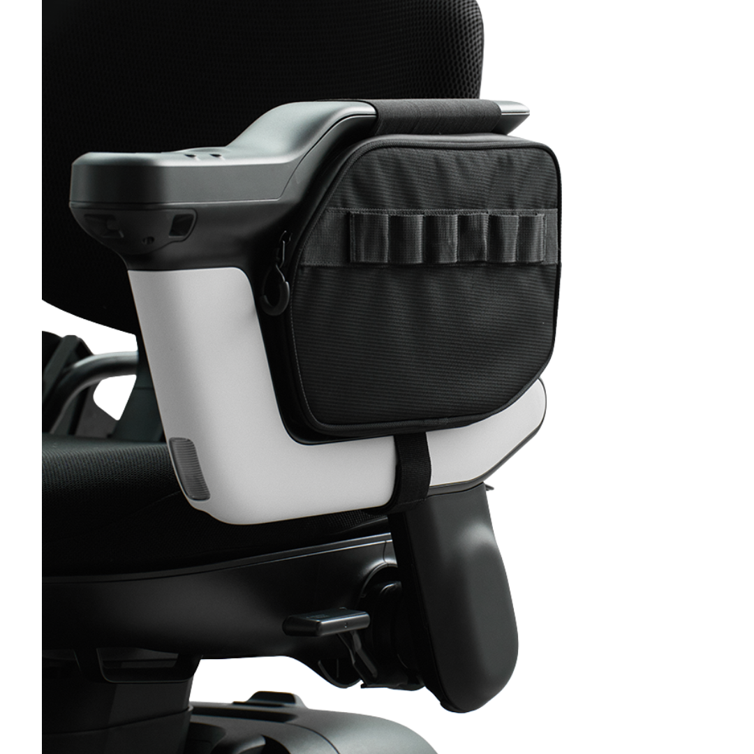 A Side Storage Bag | Strutt ev1 Compatible in black is secured to the armrest of a mobility device or wheelchair using a strap, and features a zipper closure along with external loops for organizing personal items.