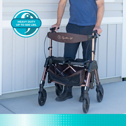 A person stands behind the Signature Life Elite Travel Rollator HD, featuring a heavy-duty brown frame, extra-wide seat, storage pouch, and a circular label that reads "Heavy Duty Up to 500 lbs," against a light gray wall and concrete ground.