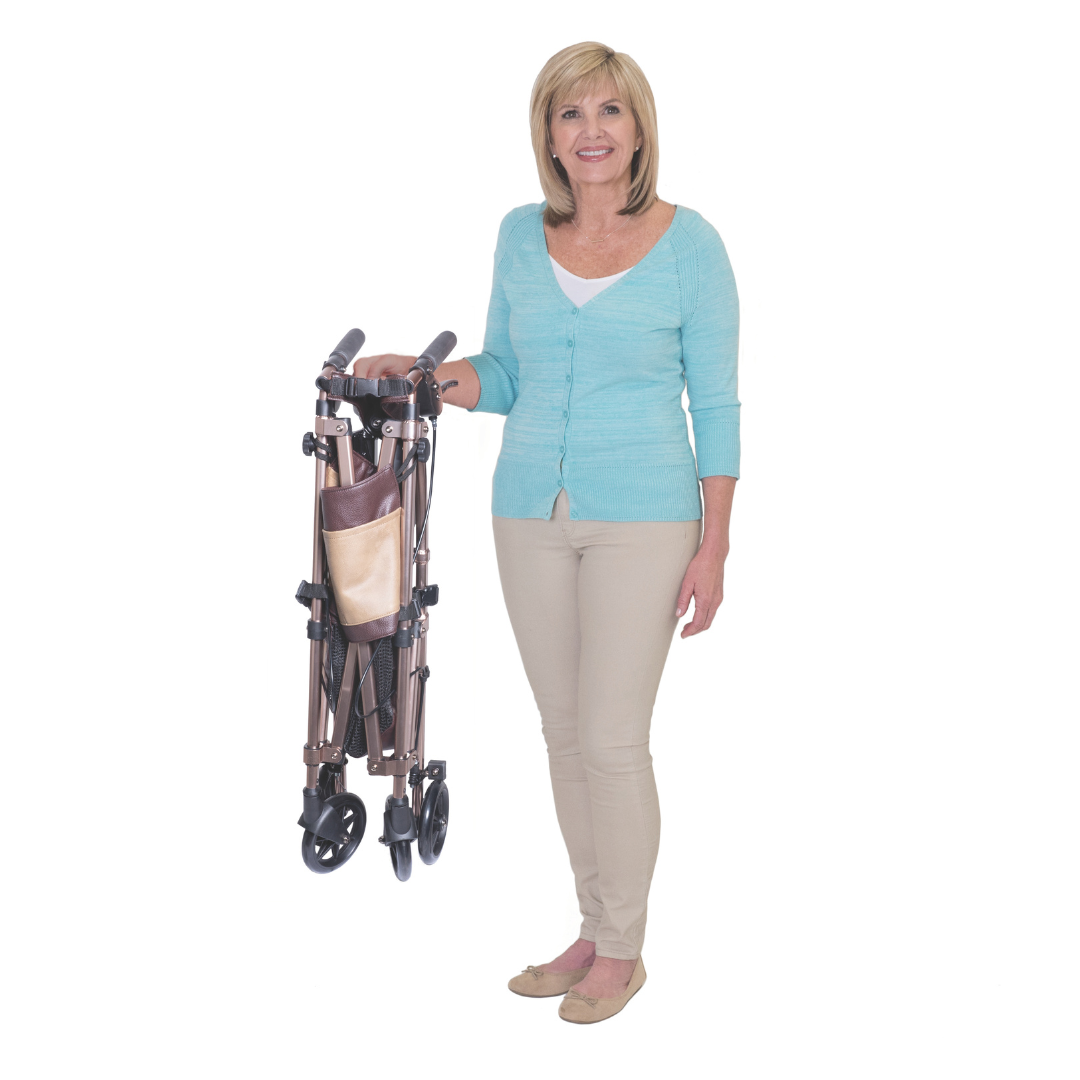 A smiling woman in a light blue cardigan and beige pants stands holding the Signature Life Elite Travel Rollator, a lightweight folding rollator with wheels and a storage pouch. The background is plain white.