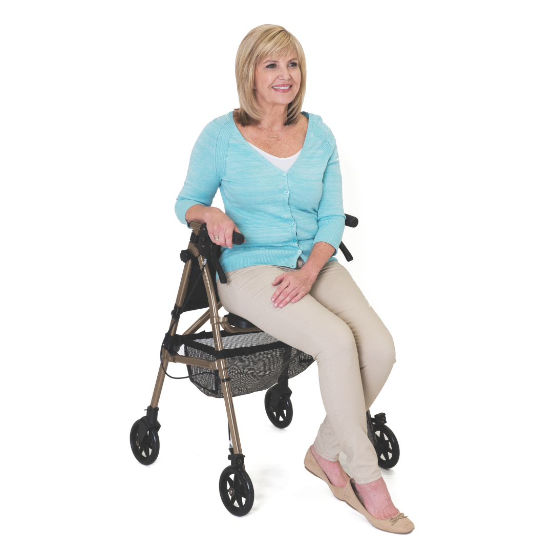 A smiling blonde woman in a light blue cardigan, beige pants, and flats sits on the Signature Life Elite Travel Rollator with a basket underneath, set against a plain white background.