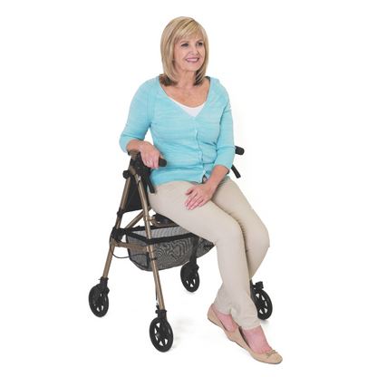 A smiling blonde woman in a light blue cardigan, beige pants, and flats sits on the Signature Life Elite Travel Rollator with a basket underneath, set against a plain white background.