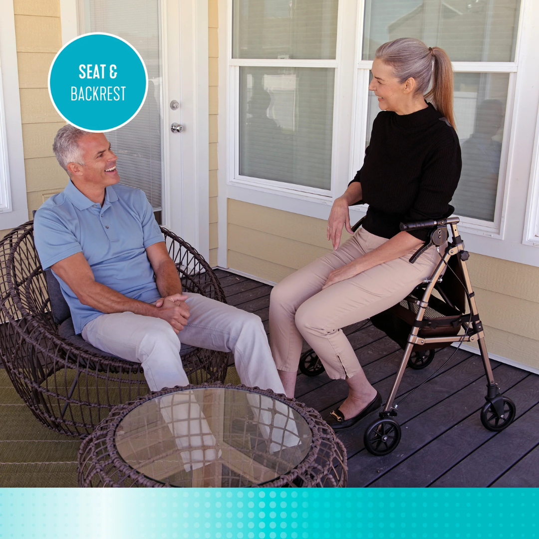 A smiling man sits in a wicker chair on a porch, chatting with a woman who uses the Signature Life Elite Travel Rollator, a lightweight rollator with a seat and backrest. Both look relaxed as they enjoy their conversation.