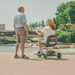 A man stands near a river while a woman sits on a Glion SnapnGO Ultralight Folding Mobility Scooter. Trees and a bridge are visible under the clear sky.