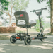 A Glion SnapnGO Ultralight Folding Mobility Scooter in black and silver, with its logo on the seat, is parked on a sunlit path. Featuring three speed settings, it sports three wheels, a backrest, and handlebars amid lush greenery and trees.