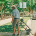 A man in a blue shirt and gray shorts strolls on a sunny sidewalk, effortlessly pulling his Glion SnapnGO Ultralight Folding Mobility Scooter. The trees sway gently, with a parked car visible in the background.