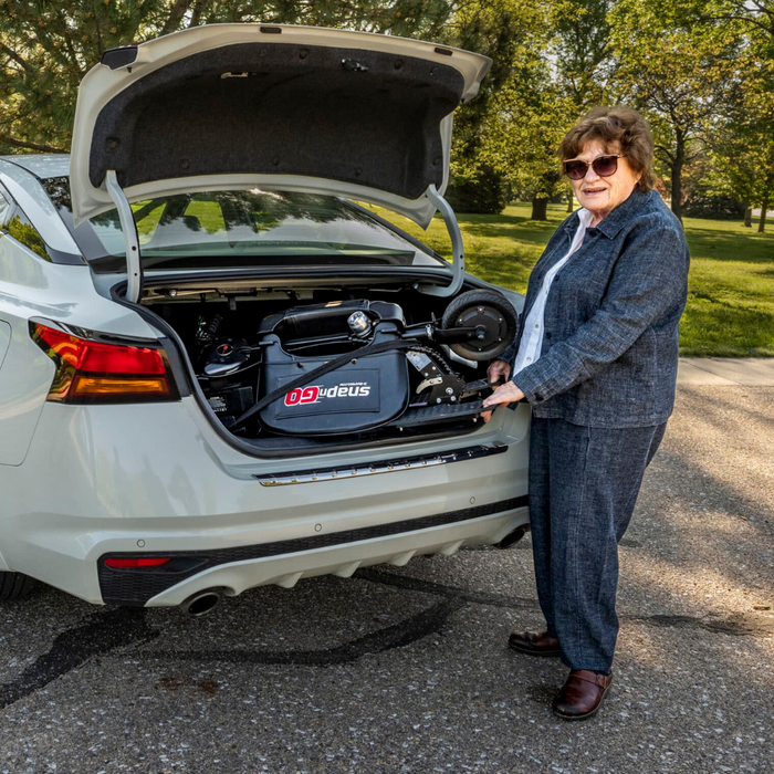 An elderly person stands beside an open car trunk, where a folded Glion SnapnGO Ultralight Folding Mobility Scooter - Airline Approved rests. Wearing sunglasses and a blue outfit, they stand on a sunlit road with trees in the background.