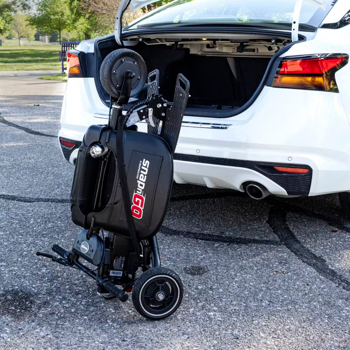 Partially unfolded near a white cars open trunk, the Glion SnapnGO Ultralight Folding Mobility Scooter flaunts its sleek black design with red accents and three speed settings in a serene parking lot surrounded by trees and grassy areas.