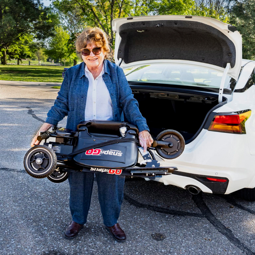 An elderly woman in sunglasses and a blue outfit stands by an open car trunk, holding her Glion SnapnGO Ultralight Folding Mobility Scooter. With three speed settings, it offers convenience and choice as she enjoys the day, with trees and a parking lot in the background.