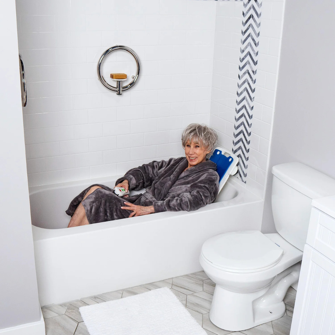An older woman in a gray robe relaxes in her bathtub using the SolutionBased BathLyft, a blue battery-powered bathtub lift chair, smiling while holding its remote control in a modern bathroom with white tile walls and a nearby toilet.