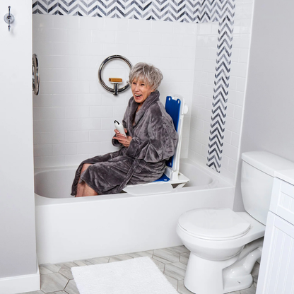 An older woman in a gray robe smiles while sitting on the SolutionBased BathLyft, a battery-powered bathtub lift chair, inside a modern white bathroom with chevron-patterned tile accents.