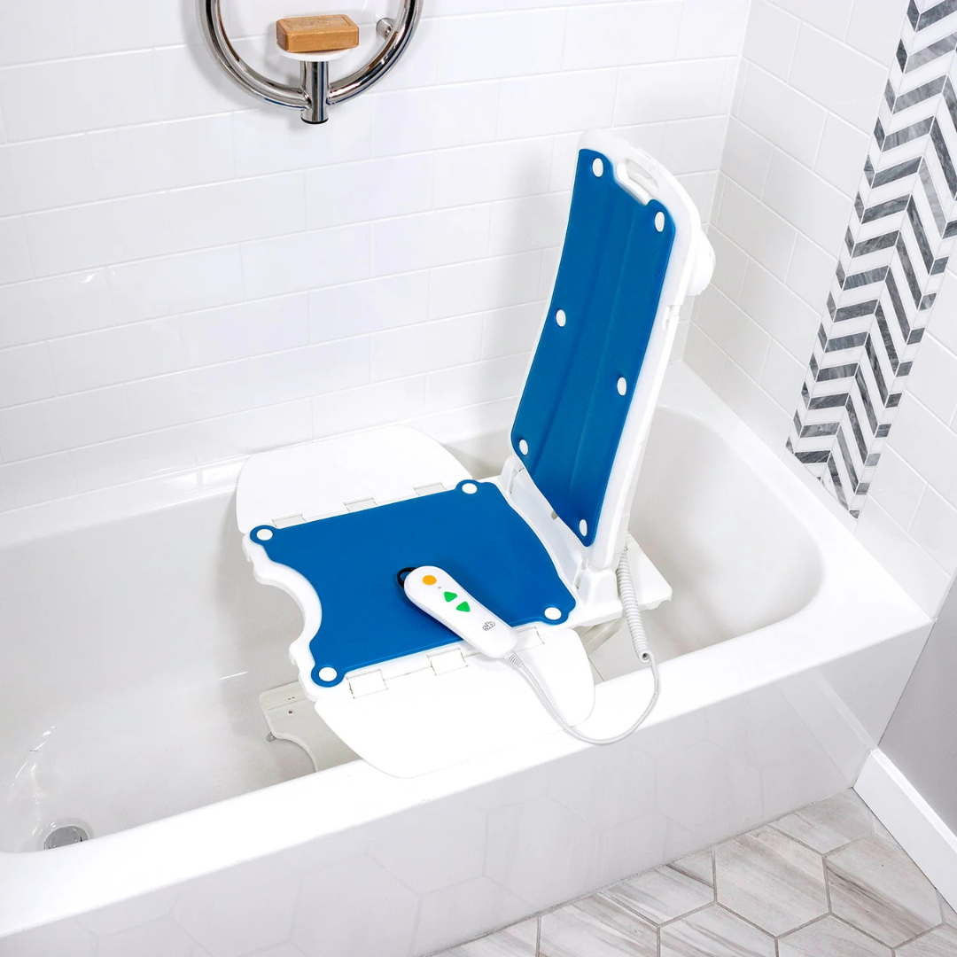 The SolutionBased BathLyft, a blue and white battery-powered bathtub lift chair with hand control, is placed inside a white tub in a tiled bathroom to help users safely get in and out of the bath.