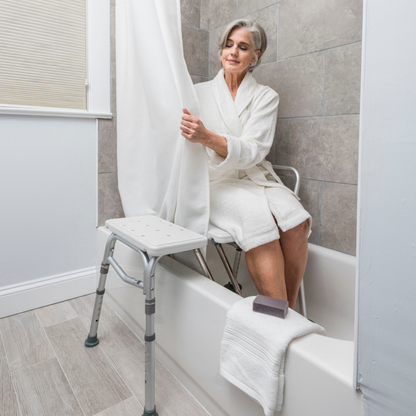 An older woman sits on the Drive Splash Defense™ Transfer Bench with Curtain Guard Protection in a bathtub, holding the shower curtain. A towel and soap rest nearby. The bathroom has gray tiles and wooden flooring for added bath safety.
