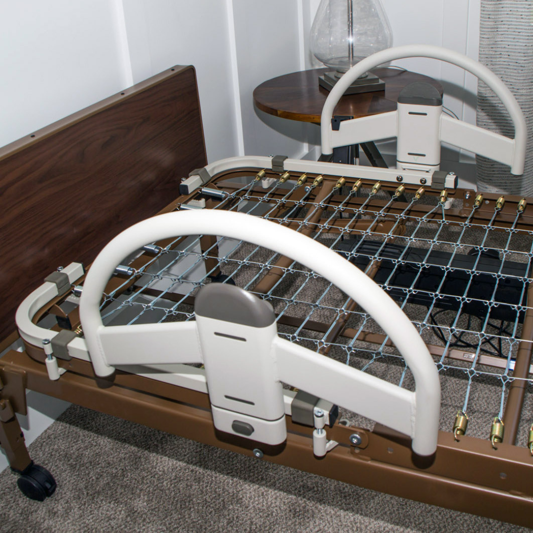 A hospital bed with Stander EZ Click LTC Bariatric Bed Handle (Set of 2), metal springs, beige rails, and a wooden headboard stands on carpet next to a round wooden side table and lamp.