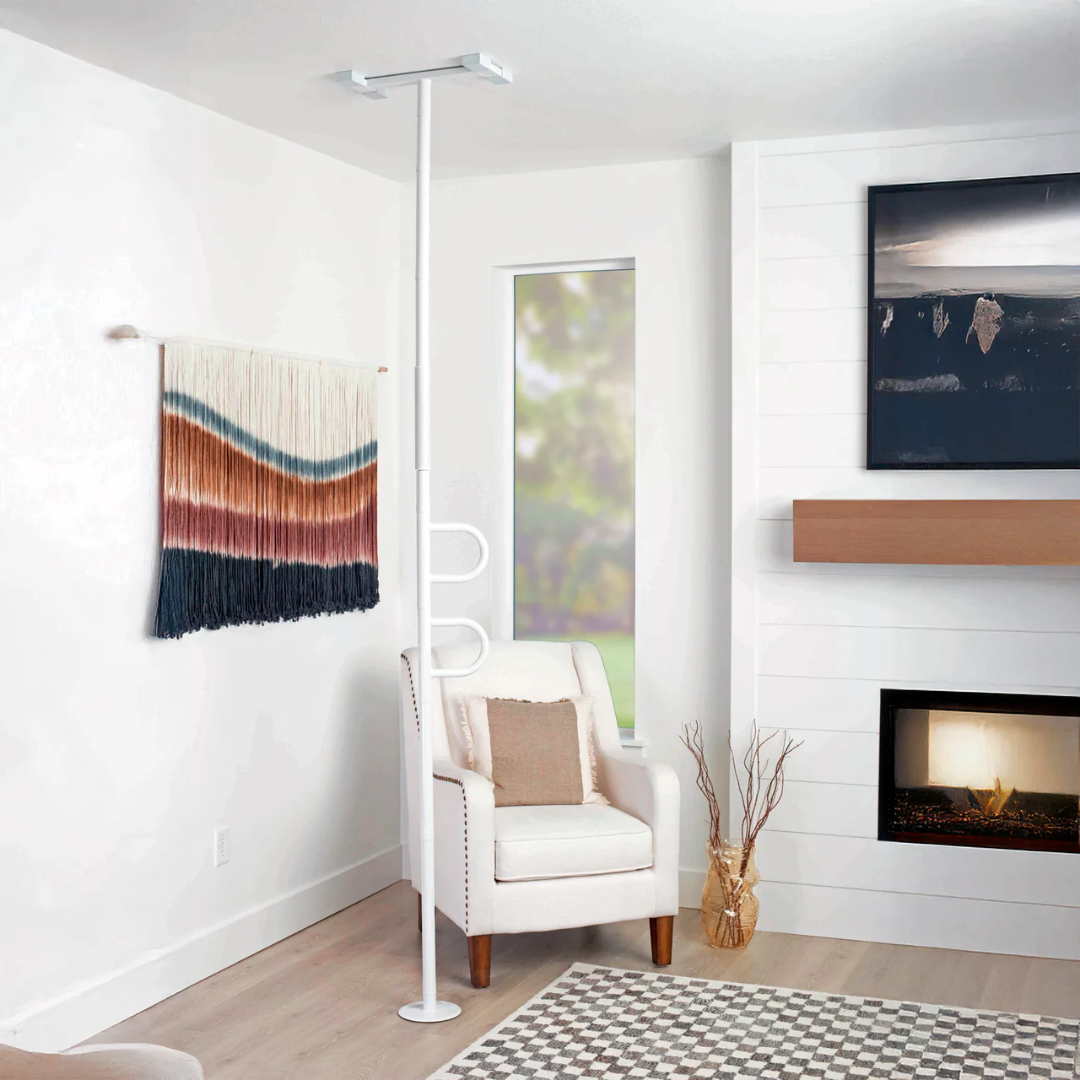 The Stander Double Handle Floor to Ceiling Grab Bar, in white with a curved handle, is installed beside a beige armchair with a pillow, near a wall hanging, window, and fireplace decorated with branches.