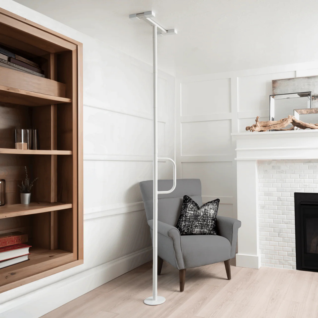 A gray armchair with a black and white pillow sits beside the Stander Wonder Pole Lite—Fall Prevention Safety Pole—in a bright, modern living room with white walls, wood flooring, built-in shelf, and white brick fireplace.