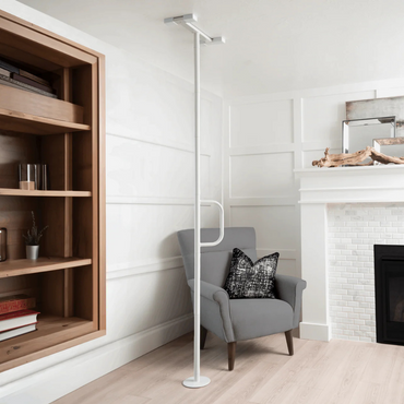 A gray armchair with a black and white pillow sits beside the Stander Wonder Pole Lite—Fall Prevention Safety Pole—in a bright, modern living room with white walls, wood flooring, built-in shelf, and white brick fireplace.