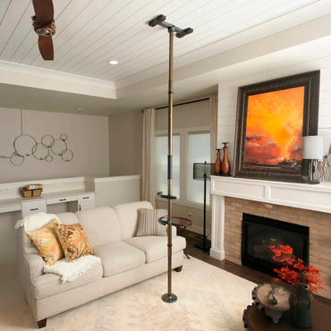 The Stander Wonder Pole Premium Fall Prevention Pole, a tension-mounted grab bar with hand grips, is installed from floor to ceiling in a living room near a beige sofa and a fireplace adorned with artwork and decorative mantel items.