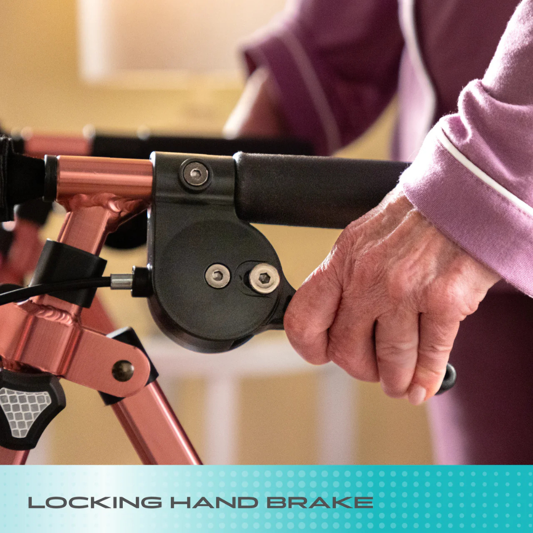Close-up of an elderly hand gripping the locking hand brake on a rose-colored Stander Wonder Rollator Plus. The person wears a purple long-sleeve top with white trim. Text at the bottom: Locking hand brake.