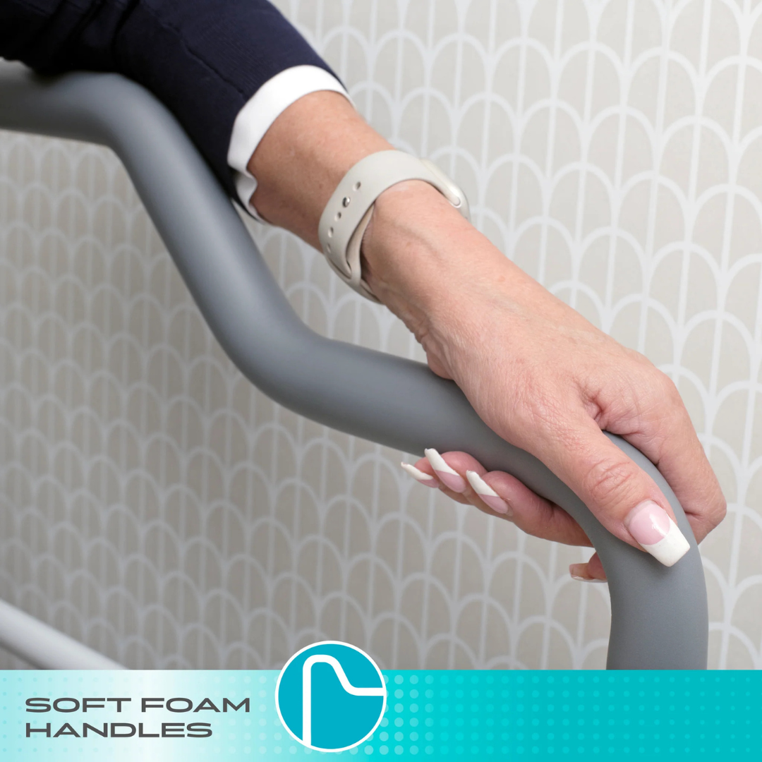A close-up shows a hand gripping the soft foam handlebar of the Stander Wonder Toilet Safety Rails & Standing Aid for bathroom stability, with geometric wallpaper in the background. A label at the bottom reads SOFT FOAM HANDLES.