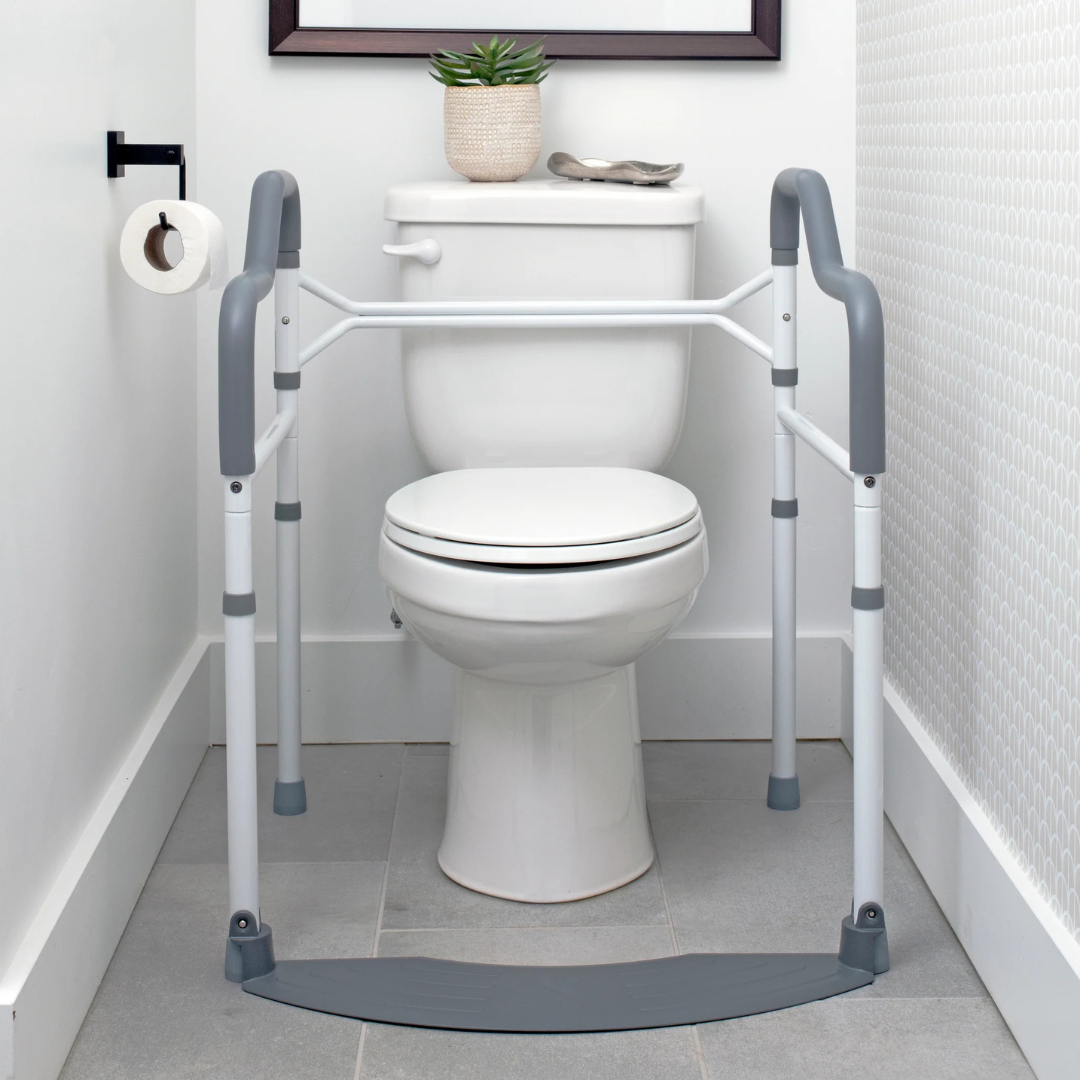 A white toilet features Stander Wonder Toilet Safety Rails & Standing Aid on both sides for stability. A toilet paper roll hangs on the left wall, and a small potted plant sits atop the tank.