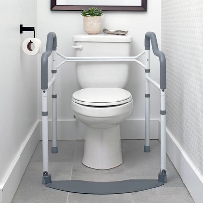 A white toilet features Stander Wonder Toilet Safety Rails & Standing Aid on both sides for stability. A toilet paper roll hangs on the left wall, and a small potted plant sits atop the tank.