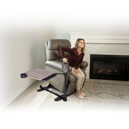 A woman uses the Stander Wonder Tray Premium - XL TV Tray & Standing Aid, attached to her lift chair, to help herself stand. The adjustable tray with a cup holder is also connected. The scene is in a living room with fireplace and patterned rug.