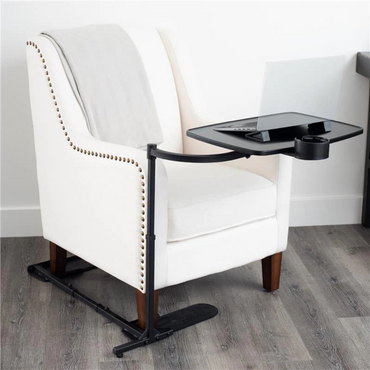 A white armchair with wooden legs and nailhead trim sits on a wooden floor. A gray blanket is draped over the chair, and a Stander Wonder Tray XL Swivel Living Room Tray Table with a cup holder is attached to the side, perfect as a laptop stand.