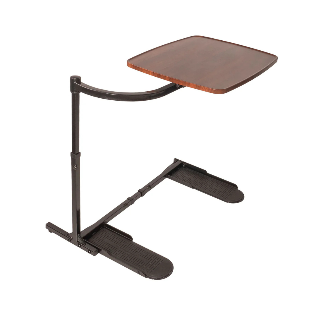 The Stander Wonder Tray is a height-adjustable, swivel tray table with a brown wooden top and black metal frame, designed for use beside a bed or chair. Its supportive base slides under furniture for added stability.
