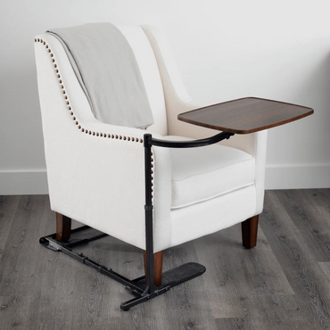 A white upholstered armchair with wooden legs holds a gray blanket and features the Stander Wonder Tray - Swivel Living Room Tray Table extending over the seat. The chair is on a gray wood floor set against a white wall.