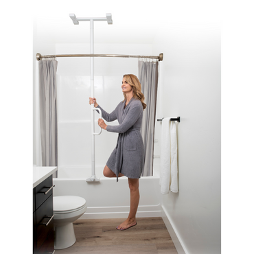 A woman in a gray robe smiles as she holds the Stander Wonder Bathtub Pole Lite next to a tub in a bathroom featuring gray curtains, white walls, a towel rack, and toilet—showcasing safe entry and exit with one foot on the tub edge.