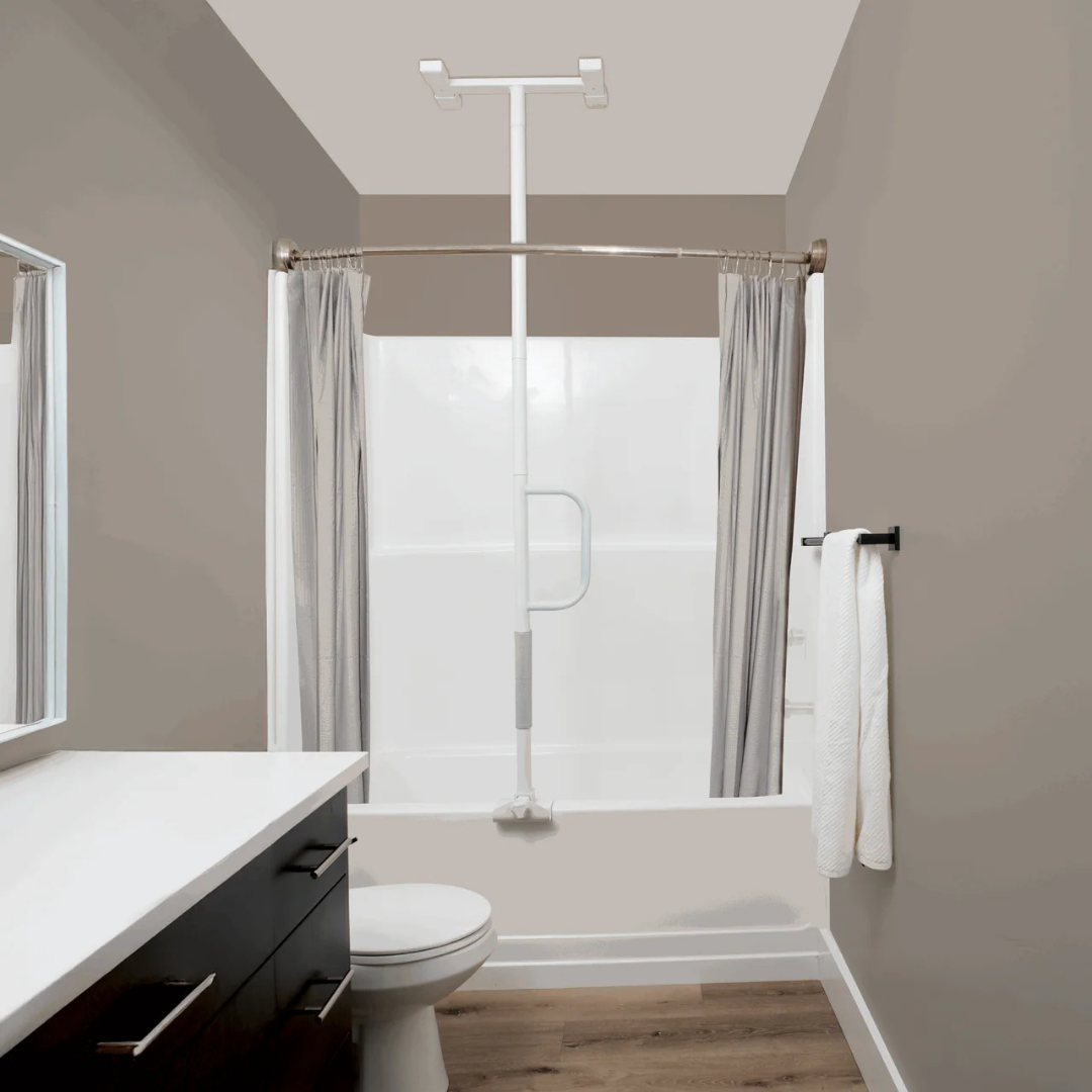Modern bathroom with a white bathtub and shower, gray shower curtain, Stander Wonder Bathtub Pole Lite for safety, white towel on a rack, toilet, white countertop, brown cabinets, and wood flooring.