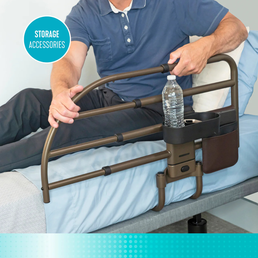 A person on a bed grips the Signature Life Freedom Click Extendable Bed Rail for fall protection, which includes storage accessories like a water bottle holder and pocket, beside blue and white bedding.