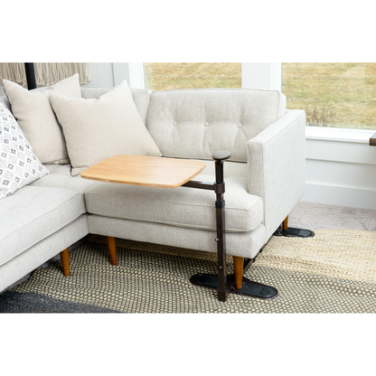 A Stander Omni Tray – TV Tray & Standing Aid, featuring a wooden swivel table and black adjustable support arm, is attached to a light-colored sectional sofa with beige cushions in a bright living room.