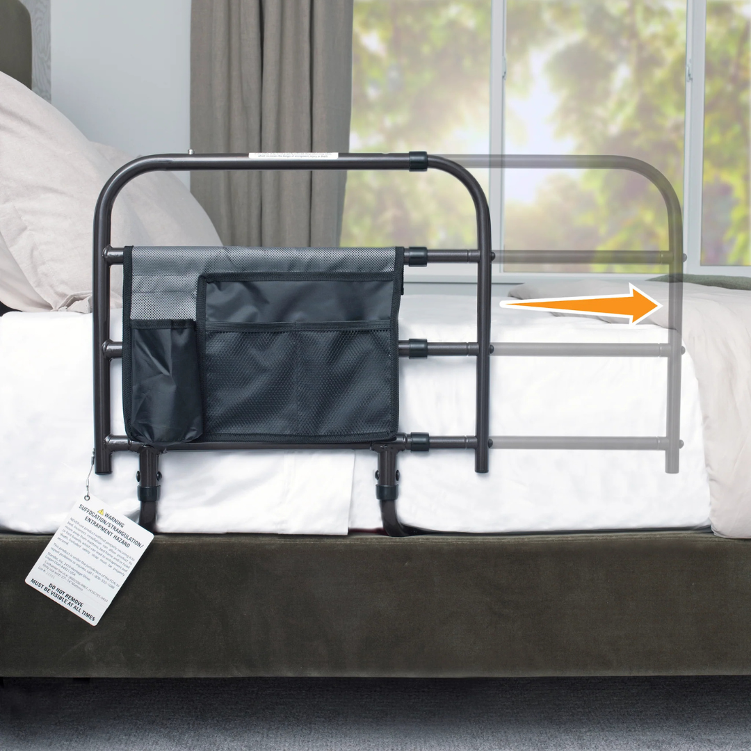 A Stander Wonder EZ Adjust Bed Rail - Extendable in black, featuring storage pockets, is attached to a bed. An orange arrow indicates the rail’s ability to extend outward, showcasing its adjustable length. A window with green trees appears in the background.